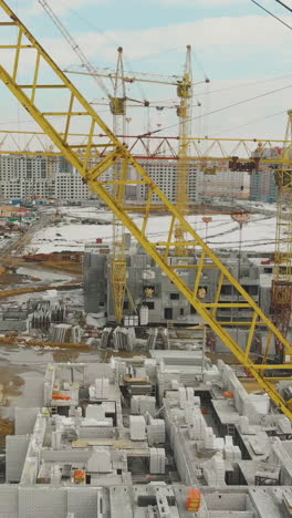 building of new dwelling complex with yellow construction cranes against snowy field and residential houses aerial view
