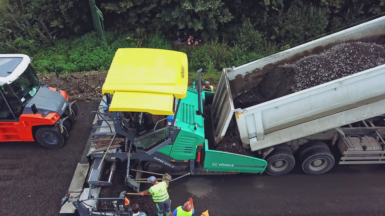 A process of laying asphalt by repairmen using an asphalt paver, a vibro-roller and a truck on a highway between cities. Aerial view.