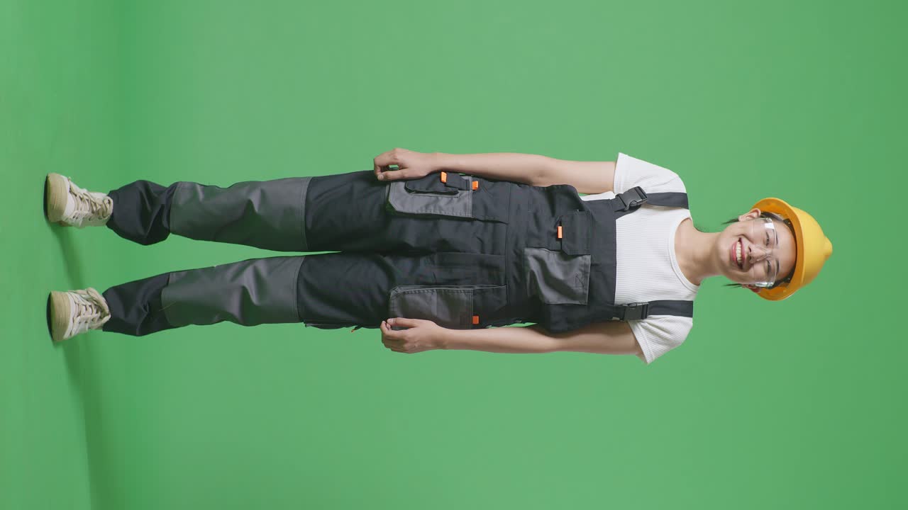 cuerpo lleno de mujer asiática trabajadora con gafas de protección y casco de seguridad de pie y sonriendo a la cámara en el estudio de fondo de pantalla verde