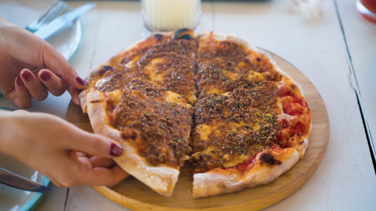 Primo piano al rallentatore di una donna con unghie curate che prende una fetta di pizza rustica con carne macinata da un tagliere di legno a un tavolo da pranzo di un ristorante informale.