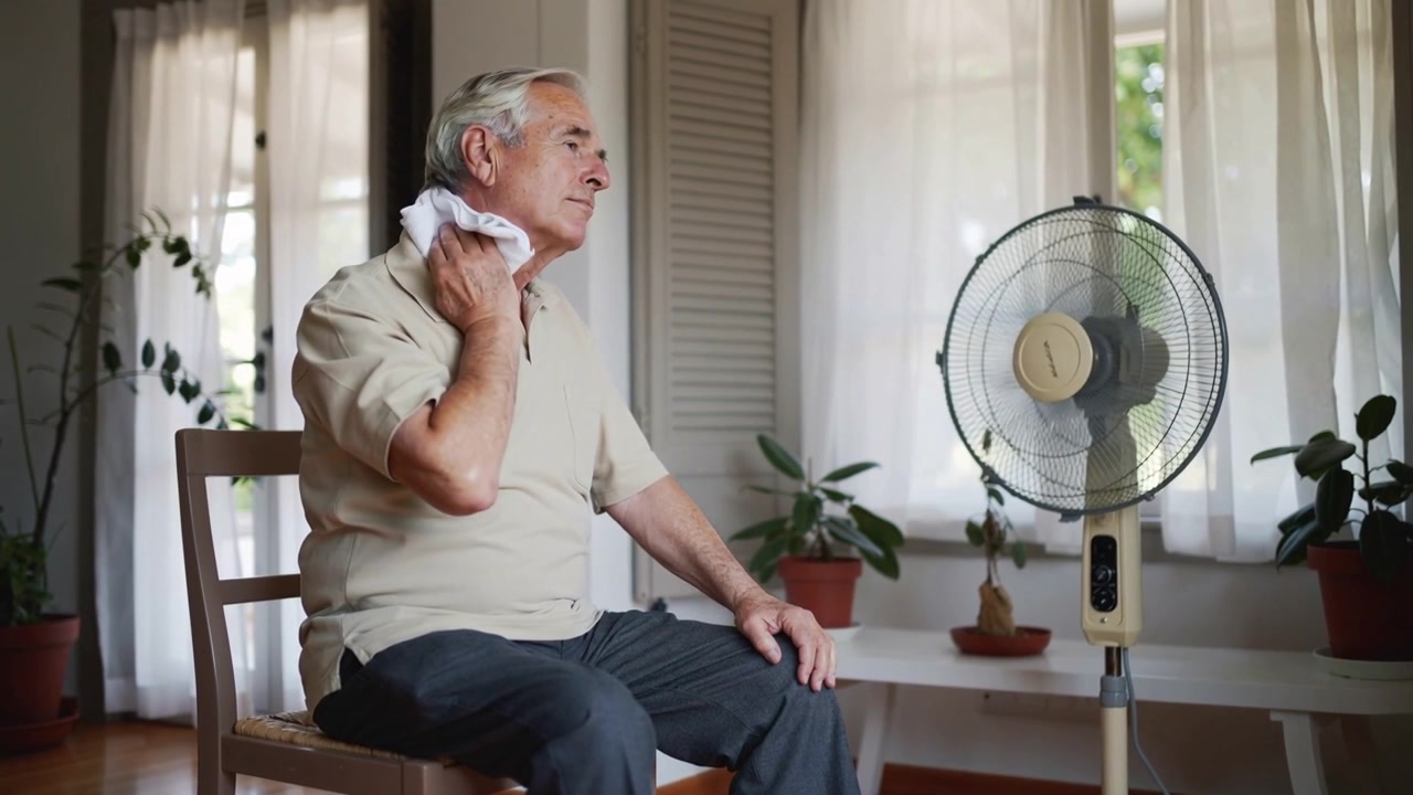 Feeling heat, wiping white cloth, senior in beige polo cooling neck facing fan in living room