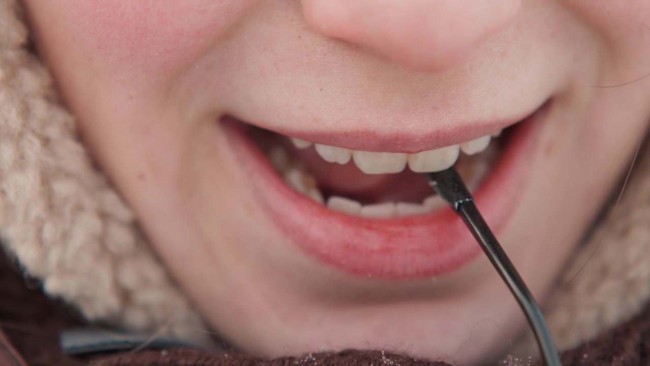 Tight horizontal close view of lower face with lips and teeth clamping thin cable or wire, tongue and jaw visible during brief biting movement, subtle playful or focused expression with winter