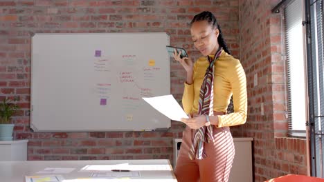 african american casual businesswoman reading document talking on smartphone in office, slow motion