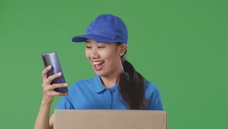 close up of asian female courier in blue uniform using smartphone while delivering a carton on green screen background in the studio