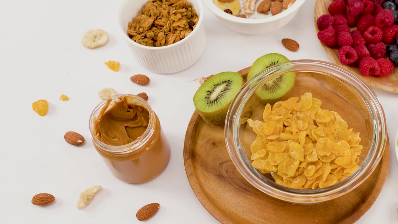 Fiocchi di mais si versano in una ciotola di vetro circondati da kiwi, noci e burro di arachidi.