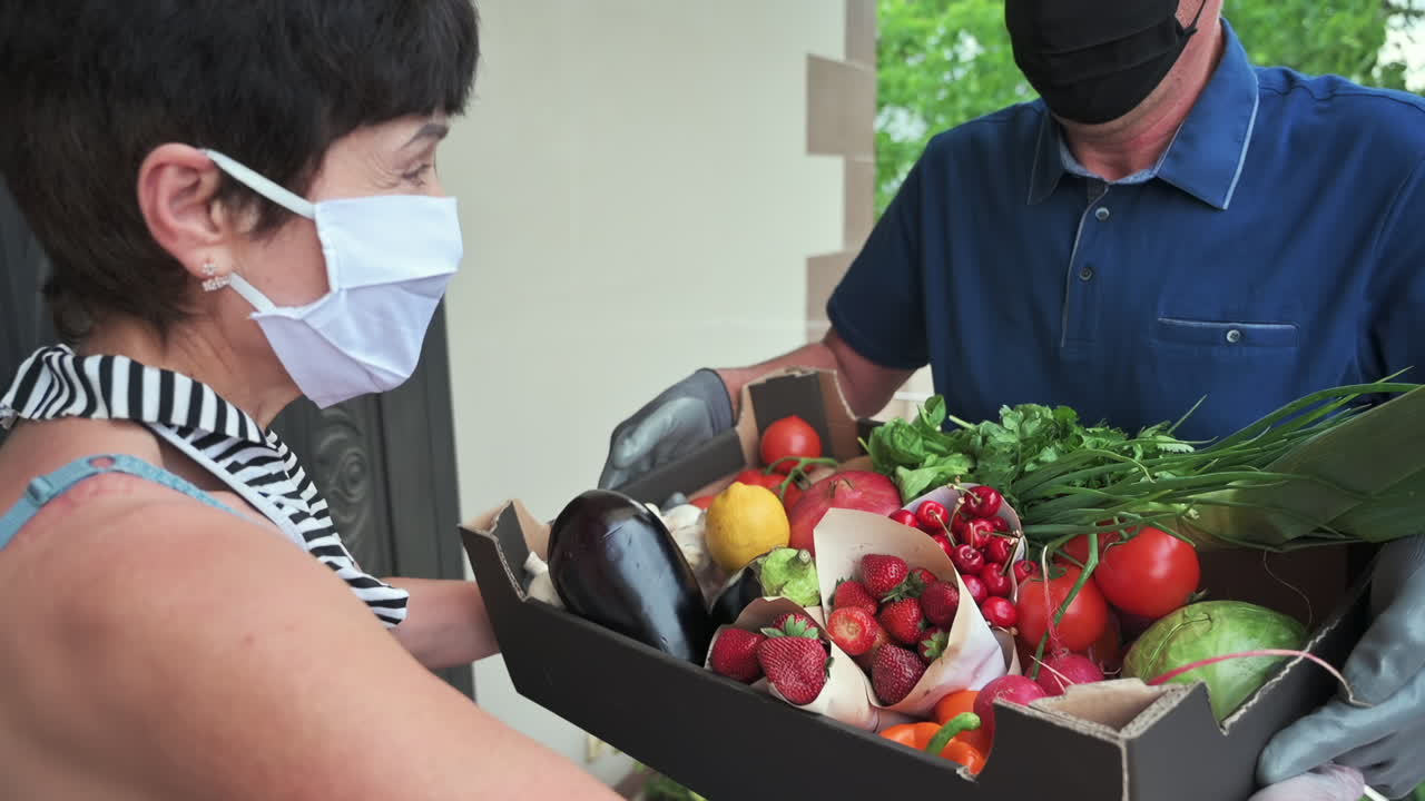 Woman receiving a box of fresh fruits and vegetables from a masked delivery man