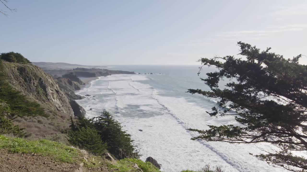 A wide-angle aerial of Big Sur’s dramatic coastline captures waves rolling toward the cliffs beneath a clear sky, framed by windswept cypress trees.