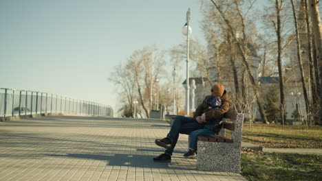 a father and son sit together on a park bench, the father resting his head on his son's head, holding him close while crossing his legs, they both dangle their legs, with a view of houses and trees