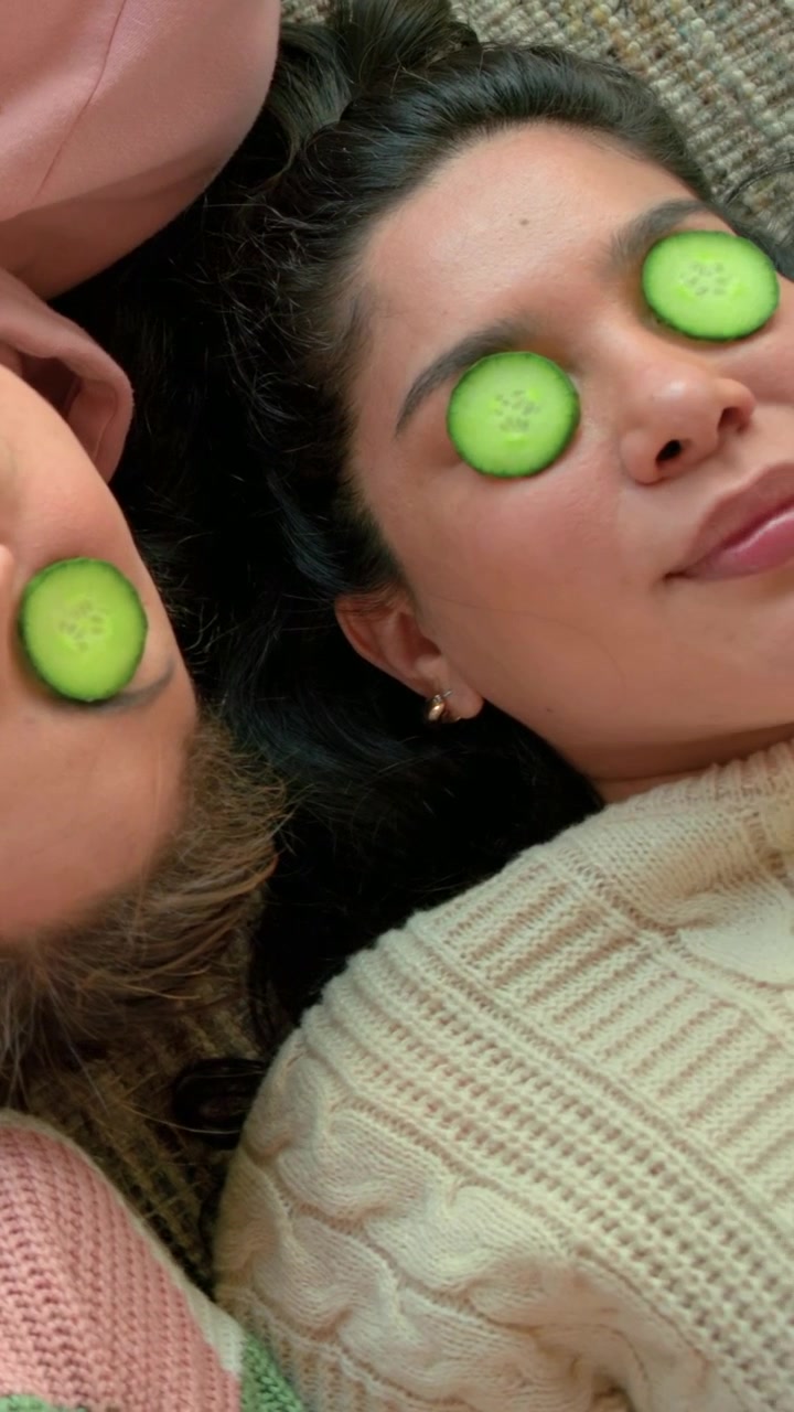 Lying two women relaxing on couch at home, wearing knit sweater, pink hoodie, cucumber slices