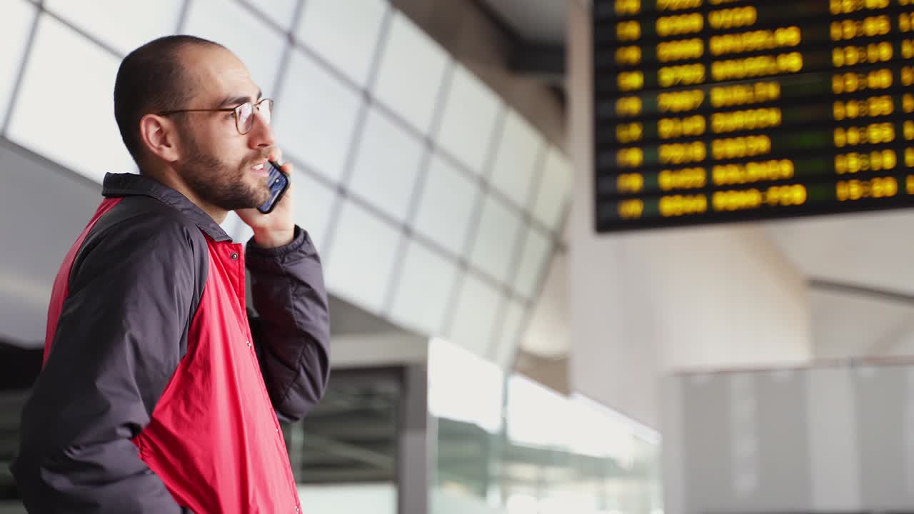 joven hablando en su teléfono inteligente por el panel del aeropuerto esperando un vuelo