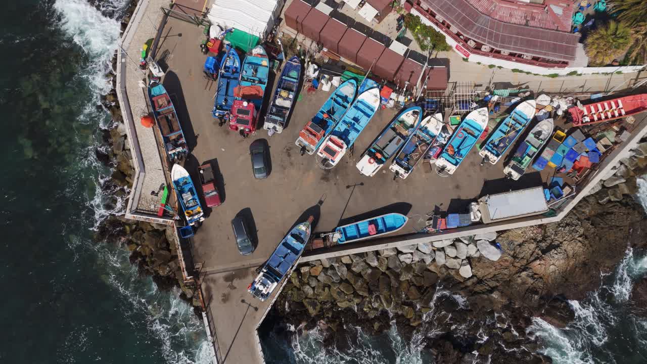 Drone shot capturing a variety of boats moored at Caleta Quintay harbour, showcasing a vibrant coastal scene and the dynamic ocean waves surrounding the pier.
