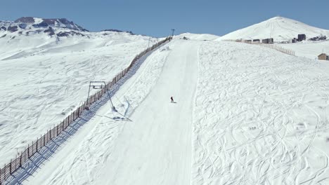 Slow-motion-aerial-view-of-a-person-with-skis-on-the-descent-of-Farellones-Park-with-a-ski-lift-on-the-side,-Chile