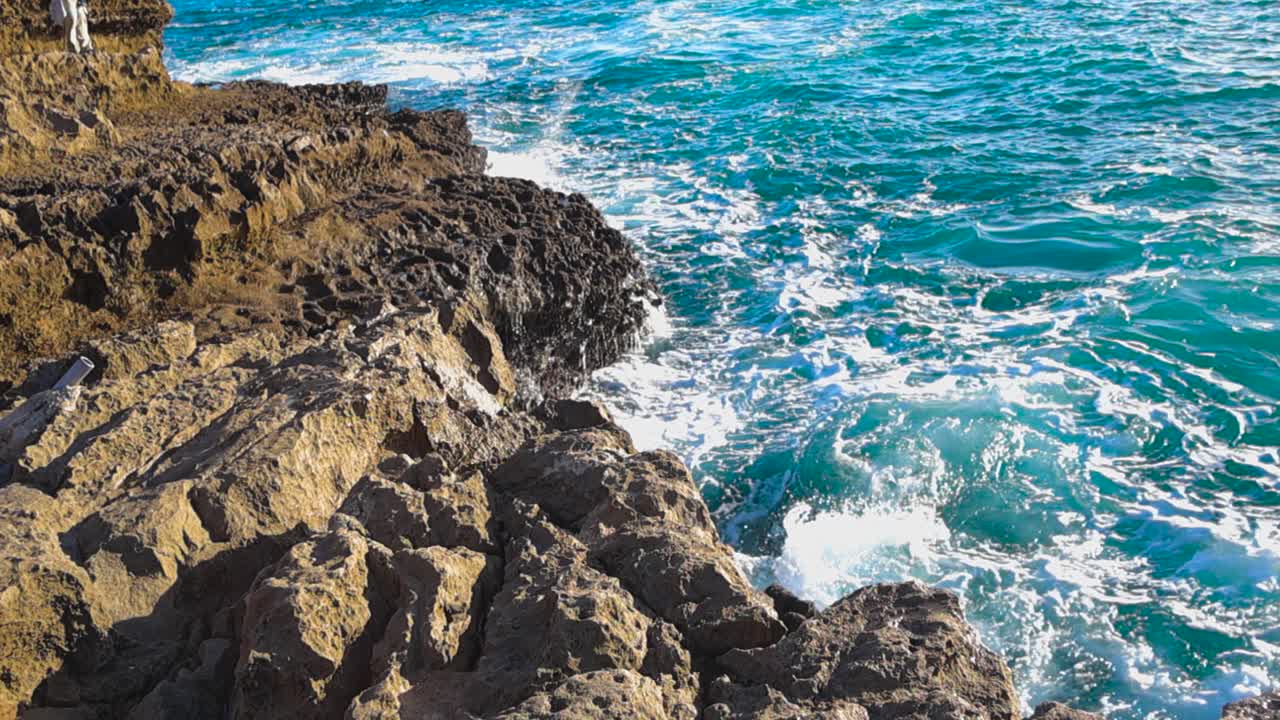 Ondas azuis batem na costa rochosa e íngreme enquanto o sol brilha sobre a água agitada e a espuma em cascata.