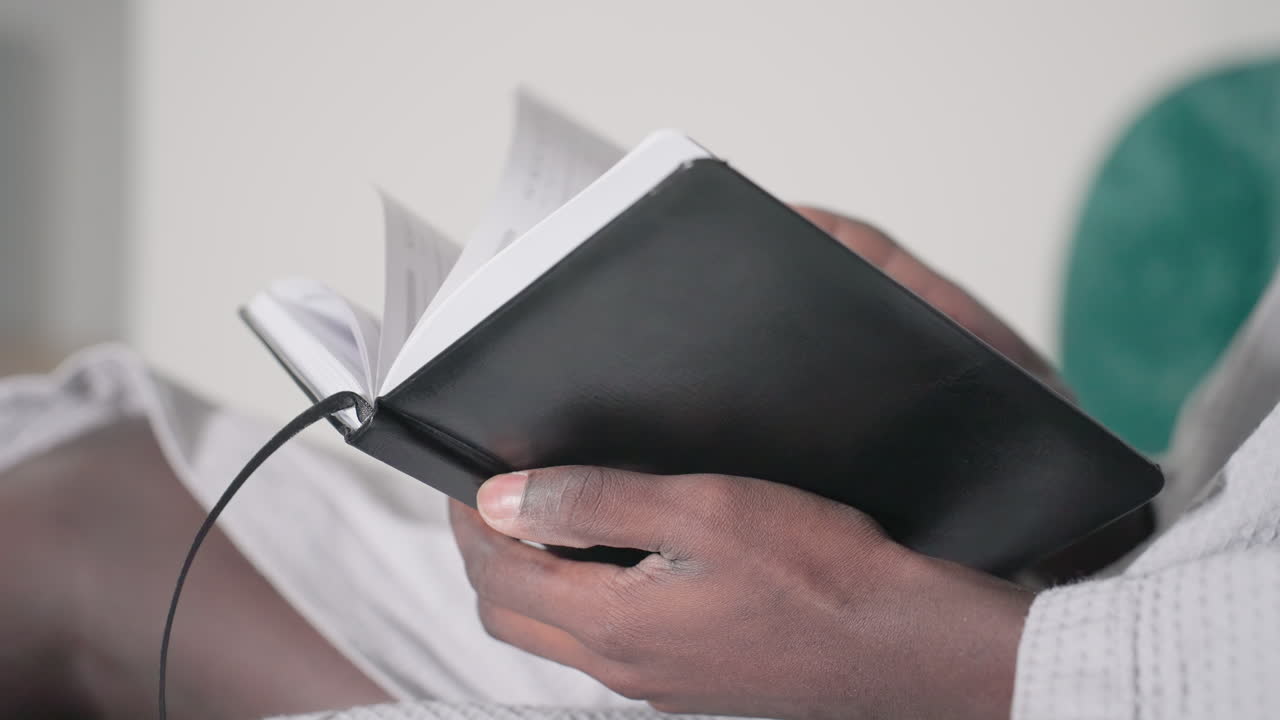 Close-up of man in white bathrobe holding open black notebook, focused on reading, soft lighting highlights hands and pages, green velvet headboard in background, peaceful indoor setting