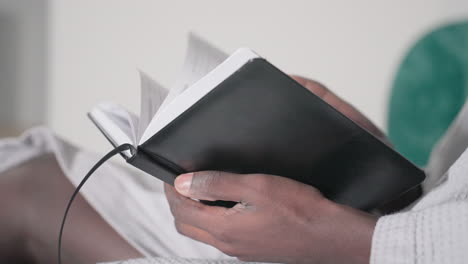 close-up of man in white bathrobe holding open black notebook, focused on reading, soft lighting highlights hands and pages, green velvet headboard in background, peaceful indoor setting