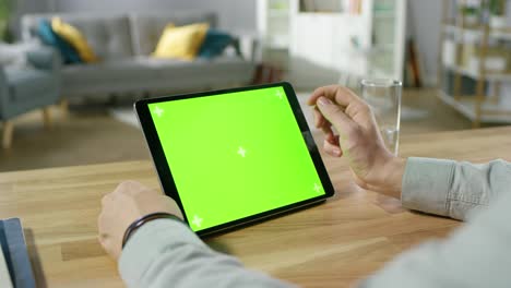 man holding and using hand gestures on green mock-up screen digital tablet computer while sitting at the desk. man buying stuff or browsing through the internet. in the background cozy living room.