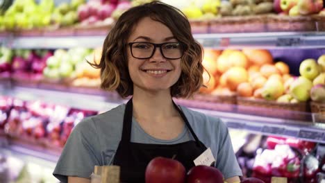 Beautiful-smiling-young-female-supermarket-employee-in-black-apron-holding-a-box-full-of-apples-in-front-of-shelf-in-supermarket