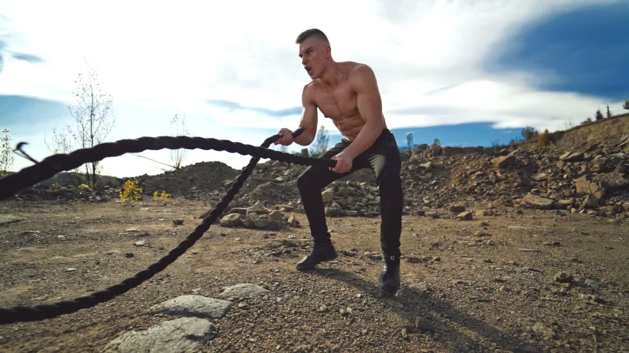 Athletic male performs battle ropes exercise at sunlight. Handsome shirtless man working with battle ropes during an outdoor fitness workout in slow motion.