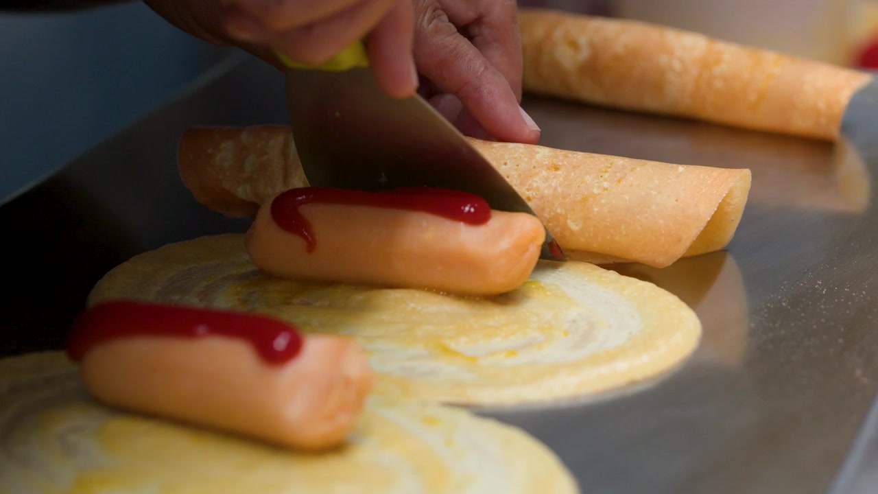 Primeiro plano das mãos enrolando panquecas finas com salsichas e ketchup numa chapa de metal.