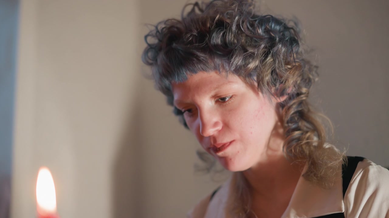 Caucasian woman blowing candle flame, intimate candlelit closeup showing curly hair and focused expression, breath creates flicker and soft amber glow, ritualistic atmosphere suggests divination