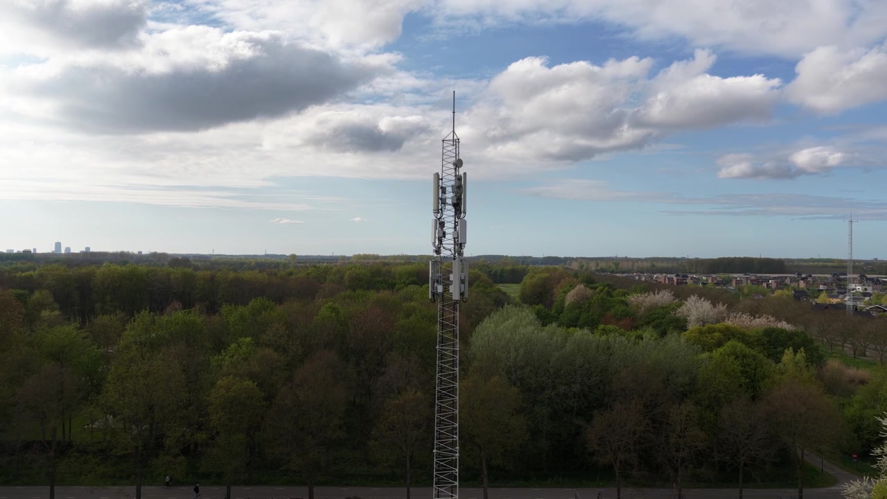 Descending aerial shot alongside a tall telecommunication tower revealing its lattice steel structure, panels, and antenna against a backdrop of a lush green forest and a partly cloudy sky