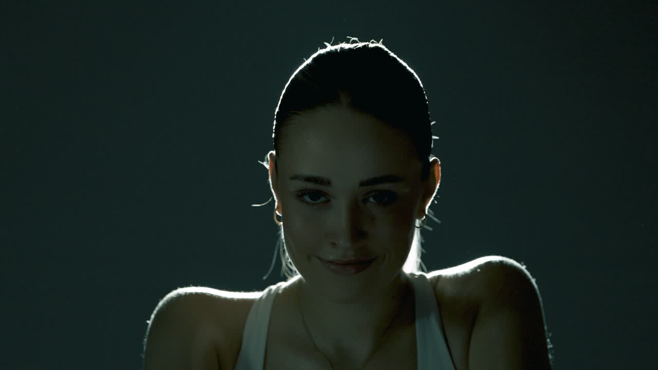 Frontal closeup of a woman cycling indoors, gazing directly into the camera with a playful, seductive smile as she pedals, blending strength and charm in soft cinematic lighting and controlled motion