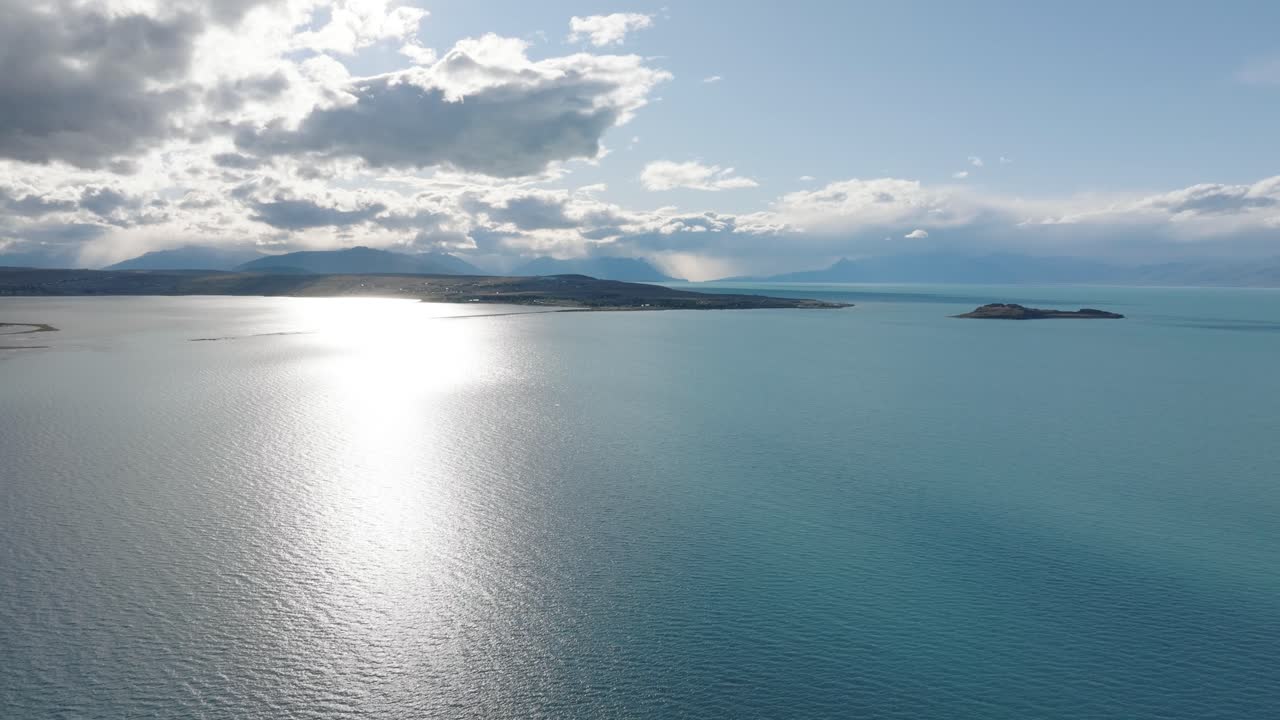 Stunning aerial view of pristine Patagonian lake with cloudy sky and distant mountains. Slow forward motion reveals nature's serene beauty with sunlight dancing on calm waters near Calafate