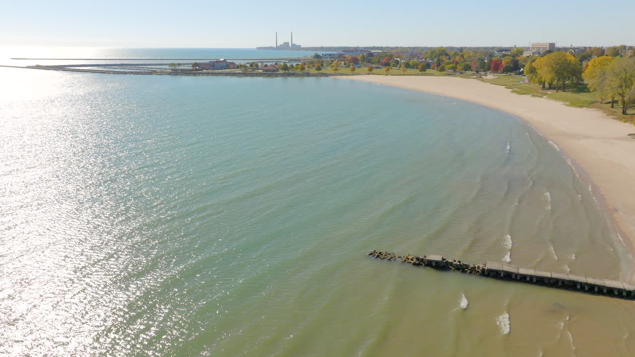 Drone aerial descending over Lake Michigan shoreline in Sheboygan, Wisconsin, showing gentle waves, colorful fall trees, and an old pier on a beautiful clear day
