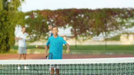 portrait of a tired teen boy coming closer to camera with racket, leaning on net and having rest after playing tennis