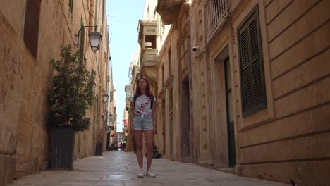 young woman looking at shop window in an alley in valetta, malta. fashion shorts clothes, sunglasses. shot in 4k. young woman standing alone in a alleyway in an older european neighborhood.