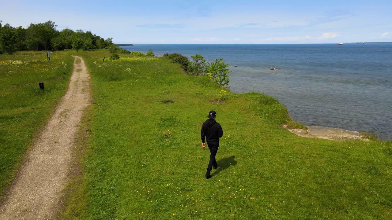 Gorgeous aerial drone view flying behind a man walking towards a tall and high altitude limestone cliff bank in Tabasalu Europe during a sunny summer day. The handsome man has black clothing on.