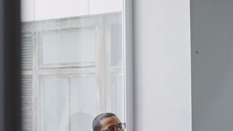 Arab-Man-With-Glasses-And-Christmas-Sweater-Sitting-In-The-Studio-Talking-On-The-Phone-With-A-Window-In-The-Background