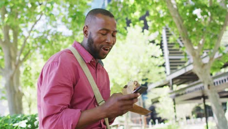 Happy-african-american-man-eating-and-using-smartphone-in-city