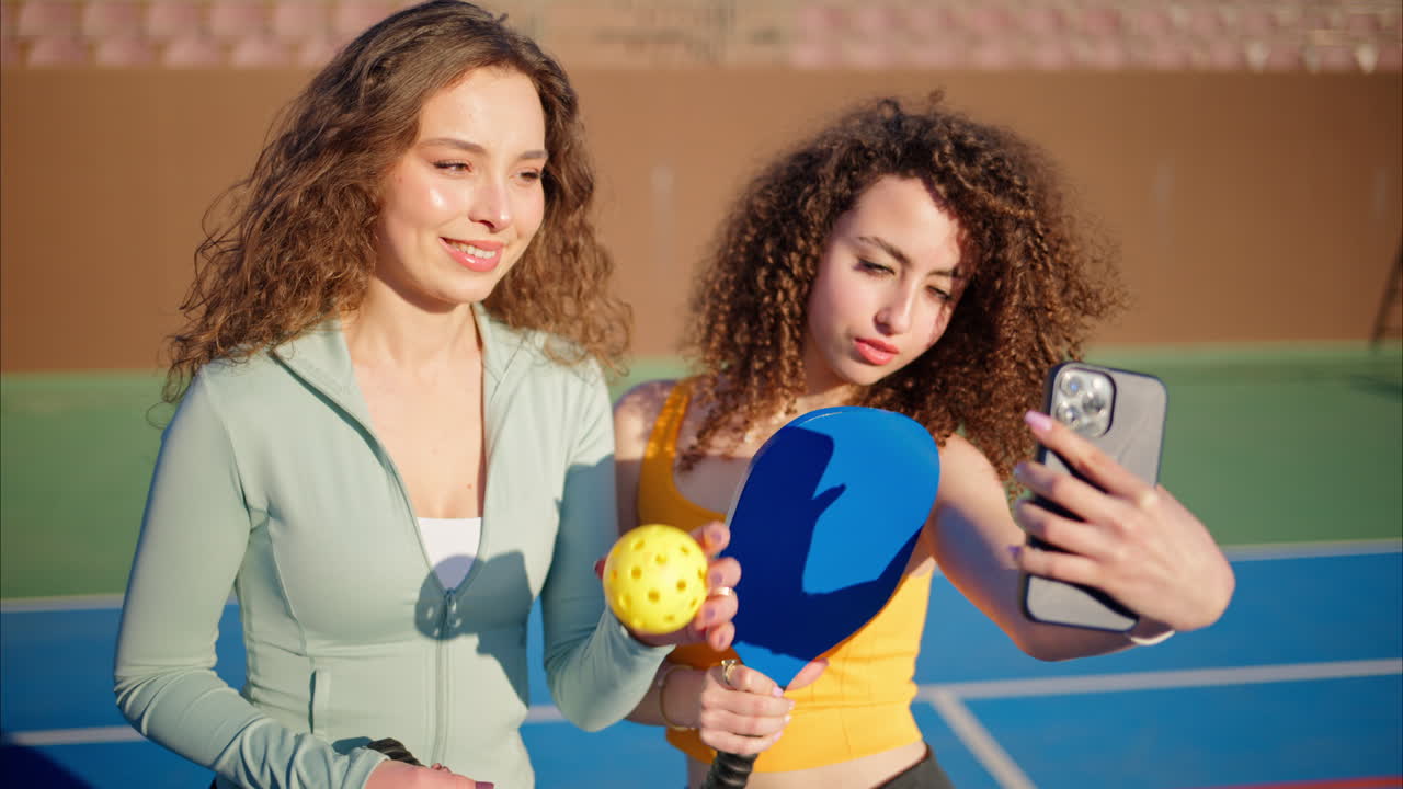 Two girls with curly hair, holding pickleball rackets smiling and talking while taking selfies on a blue court