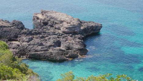 A-rock-formation-surrounded-by-turquoise-waters,-with-green-trees-in-the-foreground,-painting-a-stunning-summer-scene-in-Mallorca,-Spain