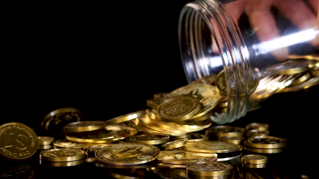 Hand pours gold-colored cryptocurrency tokens from glass jar onto black surface, dramatic lighting, close-up
