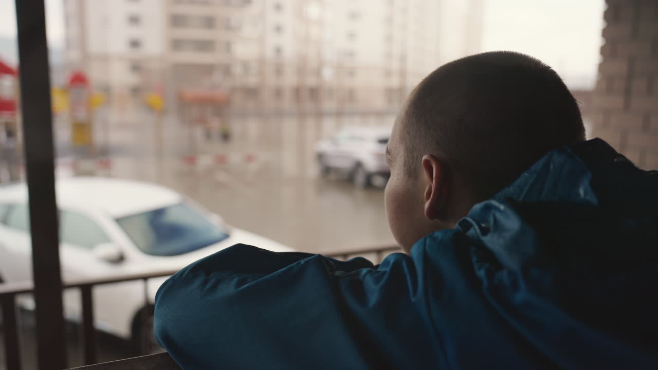 young black man scanning wet street from balcony in blue jacket, hands on railing, eyes fixed on distant cars and empty lot, resolve to return to football practice, hopeful tone with soft light