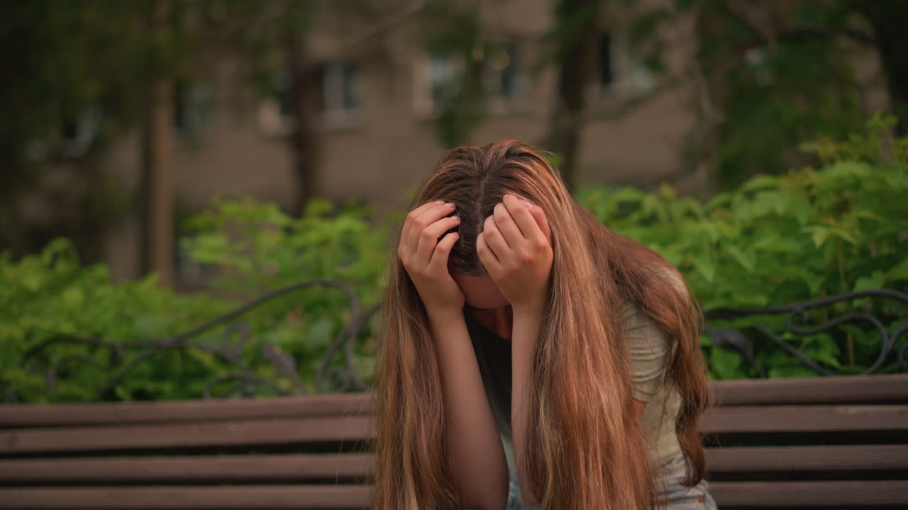 나무 벤치에 앉아있는 여자, 그녀의 얼굴을 손으로 고, 깊은 생각과 우울한 반사에 빠져, 그녀의 긴 머리카락은 그녀의 어깨에 흐르고, 울창한 초록색과 나무와 혼합됩니다.
