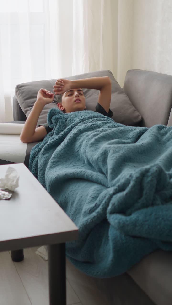 Living room with boy lying on couch covered with blanket. Glass cup and tissues on table as boy adjusts and coughs, visibly unwell and resting
