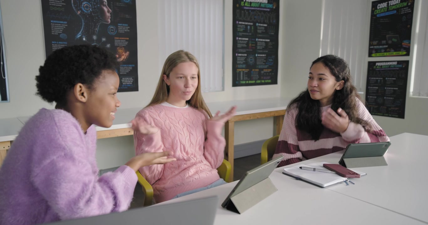 Three teen females working at study table with tablets, center student starting speaking, gesturing