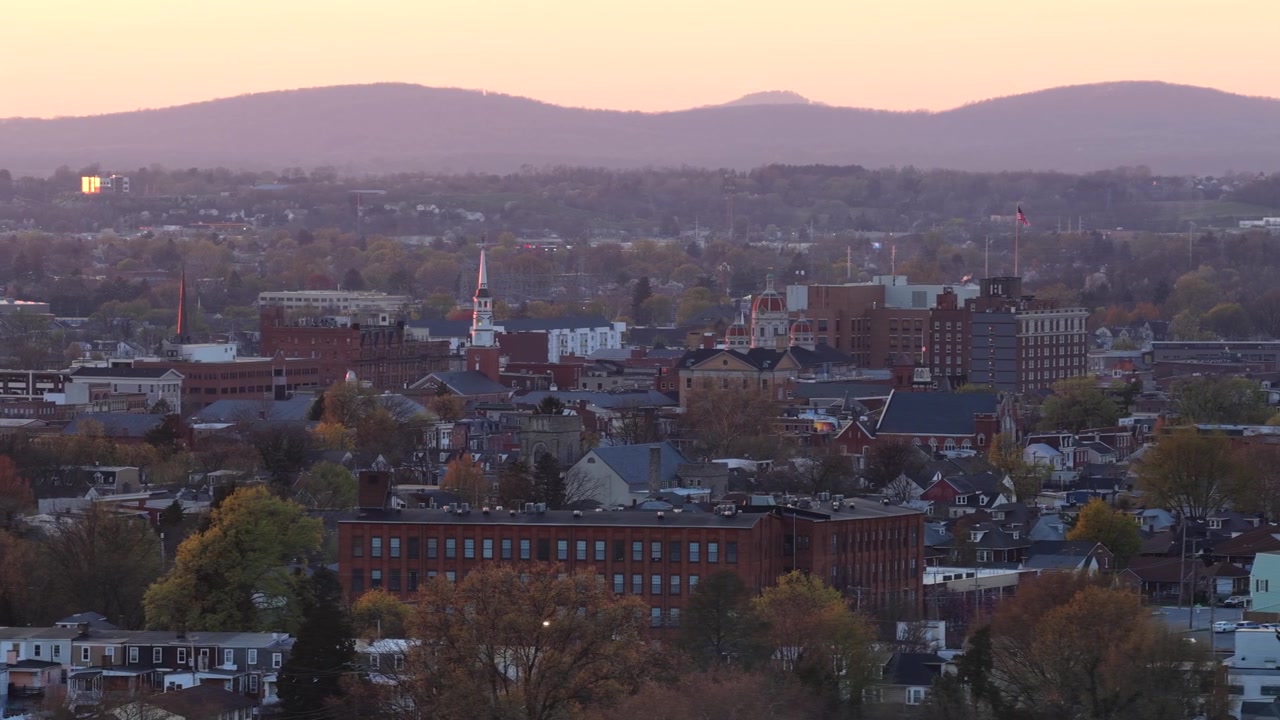 Ampia ripresa aerea del centro di York, Pennsylvania, al tramonto primaverile, con lo skyline della città, i campanili delle chiese e la bandiera americana. Montagne e una leggera nebbia si ergono sullo sfondo sotto la calda luce della sera.