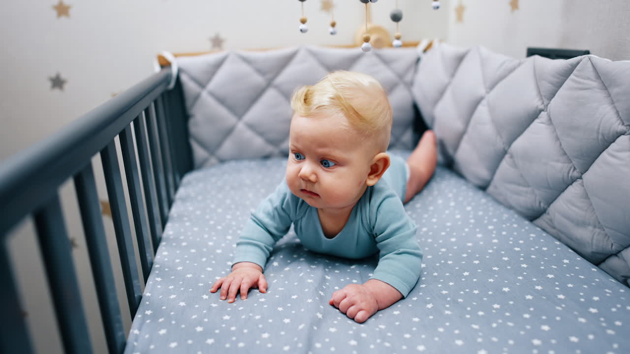 Lovely boy with plump cheeks and big blue eyes lies in the cot. Calm kid looks around peacefully.
