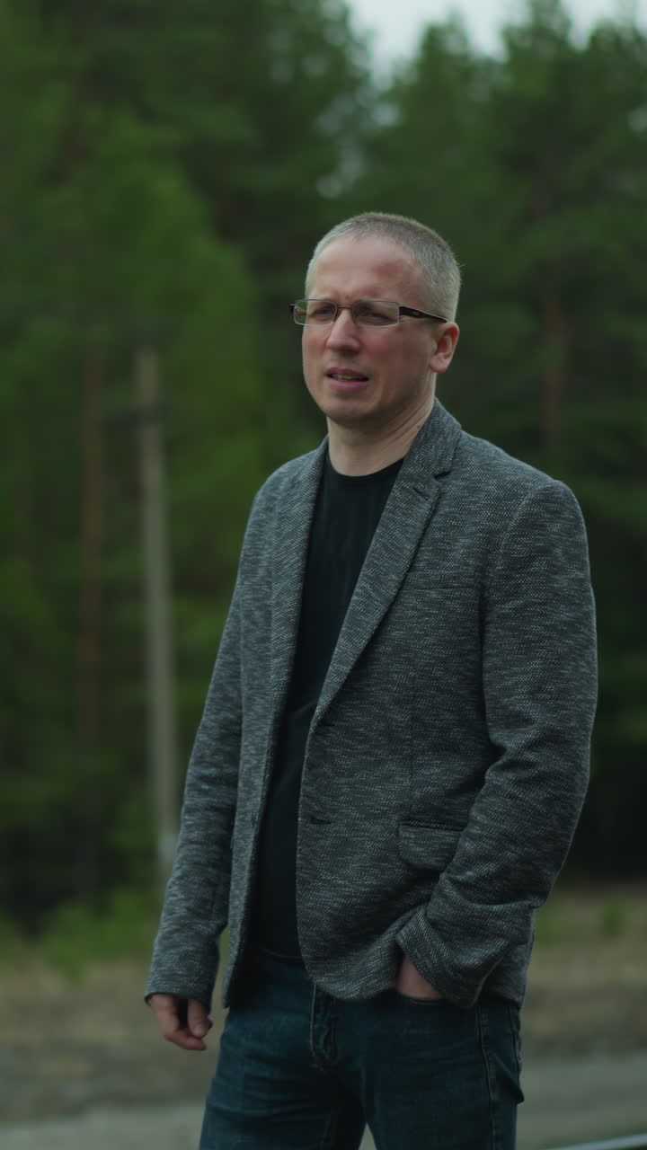 vista en primer plano de un hombre con una chaqueta gris de pie en una vía ferroviaria, poniendo sus gafas con una mano mientras mira pensativo en la distancia, con un fondo borroso