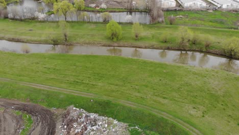 trash residues next to meadow landscape by a calm river near rural industrial structures