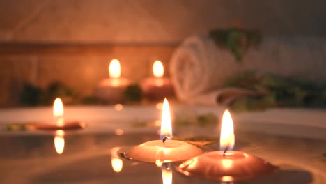 relaxing-spa-background-with-candles-floating-in-the-bath-water,-some-green-petals-and-a-towel-near-the-water-surface