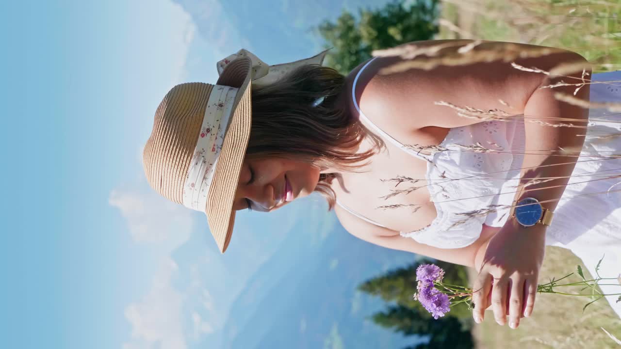 toma vertical de una mujer bonita en la naturaleza oliendo una flor púrpura durante un día soleado de verano - cordillera en el fondo, toma de retrato de cerca