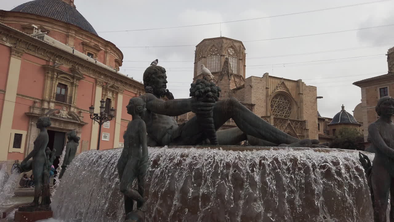 In a bustling city square, a beautiful fountain features a grand sculpture of a figure holding grapes. Water flows gracefully while pigeons rest. Surrounding buildings add to the charm of the scene