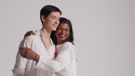 Studio-Portrait-Of-Two-Smiling-Women-Wearing-Pink-Breast-Cancer-Awareness-Ribbons-Hugging-Against-White-Background