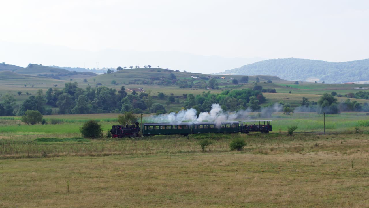 Drone. A historic steam train with passenger cars makes its journey through a vast rural landscape. The locomotive crosses golden pastures with rolling green, forested hills in the background