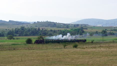 drone. a historic steam train with passenger cars makes its journey through a vast rural landscape. the locomotive crosses golden pastures with rolling green, forested hills in the background